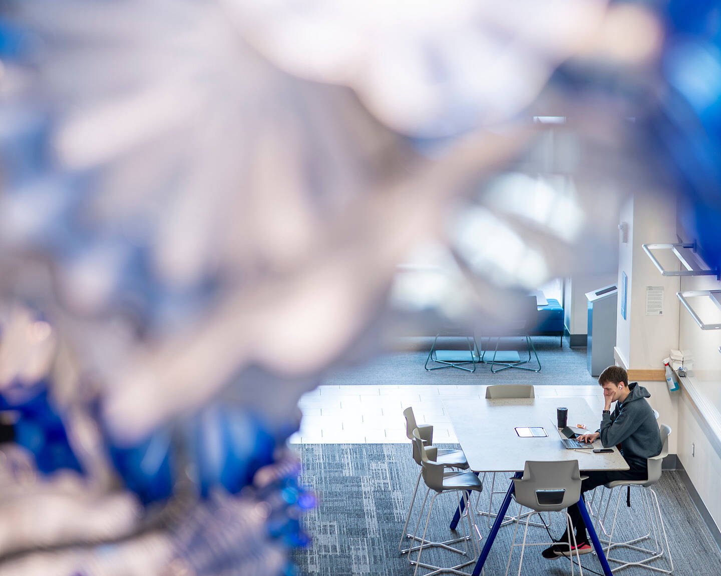 A person sits at a table in a modern, brightly lit space, working on a laptop, with blurred blue and white decorative elements in the foreground.